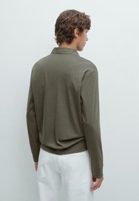 Olivgrünes Langarm-Polohemd mit umgeschlagenem Kragen, lockerer Passform und gerippten Bündchen, kombiniert mit weißen Hosen.