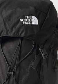 Sac à dos noir avec une surface texturée, présentant des rayures verticales. Comprend un patch logo, des bretelles réglables et une fermeture à cordon.