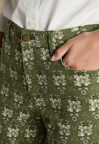 Zelené nohavice s kvetinovým vzorom majú textúrovanú látku s priliehavým strihom, kovovým gombíkom na páse a bočnými vreckami.
