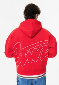 Punainen huppari, jossa suuri valkoinen graafinen logo selässä. Valmistettu pehmeästä materiaalista, rento istuvuus ja resorit hihansuissa.