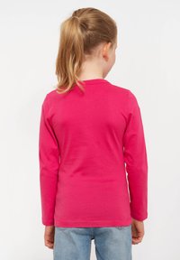 Mädchen mit blonden Pferdeschwanz, das ein leuchtend pinkes Langarmshirt und hellblaue Jeans trägt, steht mit dem Rücken zu einem weißen Hintergrund.