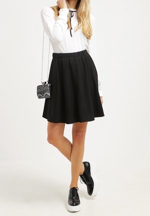 Donna che indossa una maglia a maniche lunghe bianca, gonna nera a trapezio, scarpe platform nere e una piccola borsa a tracolla nera con motivo.