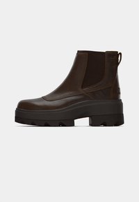 Bottine en cuir marron avec des panneaux latéraux élastiques, une languette de traction, une tige texturée, et une semelle en caoutchouc noire épaisse présentant des crampons profonds pour une meilleure adhérence.