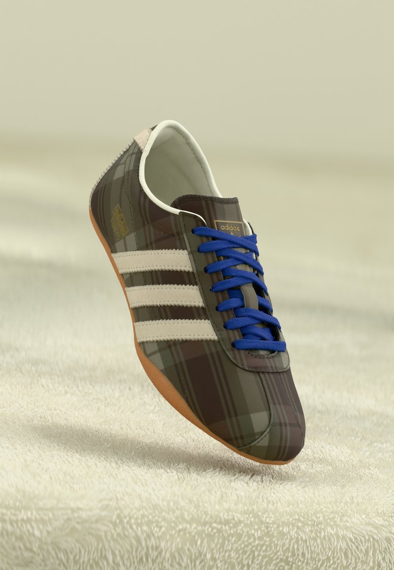 Scarpe sportive a quadri marroni con lacci blu, tre strisce bianche e una superficie liscia, dotate di una suola in gomma marrone chiaro.