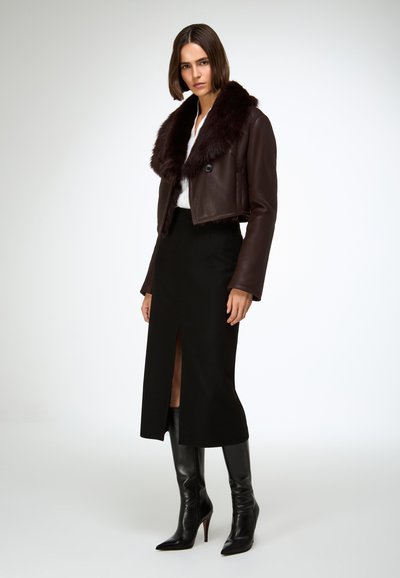 Giacca corta in pelle marrone con collo in pelliccia, abbinata a una gonna nera con spacco frontale e stivali neri alti fino al ginocchio con punta appuntita.