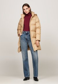 Geacă puffer bej cu glugă, cu panouri cusute, asortată cu un pulover cu guler înalt burgund și jeans bleu deschis. Botine negre completează lookul.