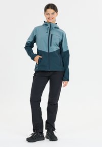 Lightweight, two-tone jacket in teal and dark navy, featuring a zippered chest pocket, adjustable hood, and elastic cuffs. Paired with black pants.