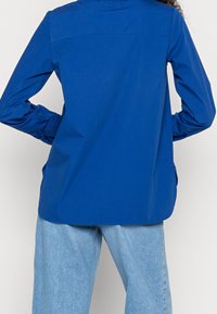 Blaues Langarmhemd mit glatter Textur, geradem Schnitt und Rückenteil-Details. Getragen mit hellblauen Jeans. Verfügt über gebogene Säume.