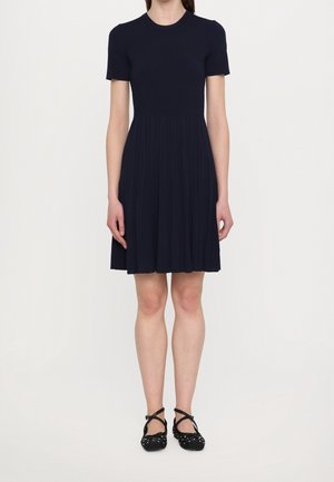 Femme portant une robe bleu marine plissée, longueur genou, manches courtes, associée à des ballerines noires cloutées avec des brides croisées.