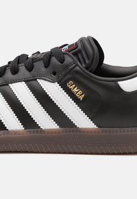 Baskets en cuir noir avec trois rayures blanches, texte "SAMBA" en doré, semelle noire texturée et col rembourré. Comprend des lacets noirs.