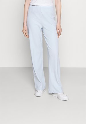 Persoon draagt lichtblauwe broek met wijde pijpen en witte sneakers, staand op een effen grijze vloer met een effen gebroken-witte achtergrond.