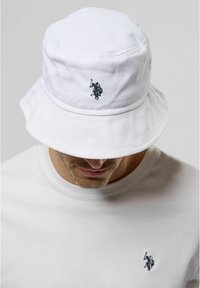 White cotton bucket hat featuring a small black embroidered logo on the front. Soft texture and wide brim. Worn with a plain white shirt.