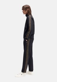 Sort træningsdragt med gyldne accentstriber, jakke med lynlås, tætsiddende bukser og hvide sneakers. Materialet ser glat ud med atletisk design.