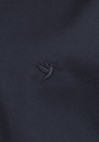 Tela azul marino con un pequeño logotipo de colibrí bordado en un tono oscuro de azul marino, que presenta una textura suave con un brillo sutil.