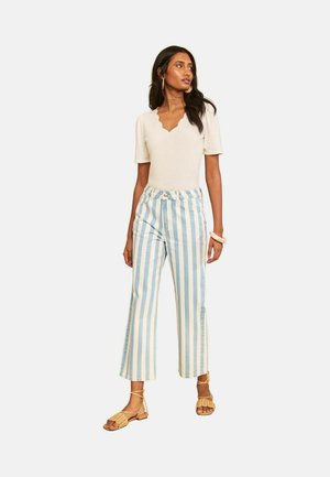 Vrouw draagt een beige top met korte mouwen met geschulpte halslijn, blauw-wit verticale gestreepte broek en beige geweven sandalen, staand op een witte achtergrond.