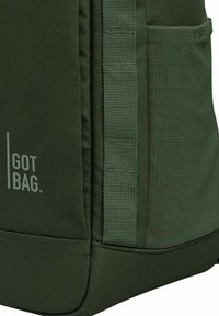 Sac à dos en tissu vert avec une surface texturée, poche latérale, et logo "GOT BAG." Caractéristiques : coutures durables et sangles renforcées.