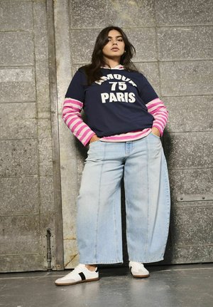 Kvinde i lyseblå vide bukser, hvide sneakers, navy skjorte med "75 Paris" over en lyserød og hvid stribet langærmet bluse, stående mod en metalvæg.