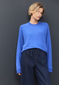Top blu a maniche lunghe con scollo rotondo e polsini a coste, abbinato a pantaloni di denim scuro a vita alta con cuciture visibili e vita elasticizzata.