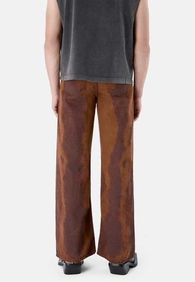 Person trägt braune Jeans mit weitem Bein und dunkleren vertikalen Flecken, schwarze Schuhe mit quadratischer Spitze und ein ärmelloses dunkelgraues Oberteil, von hinten gesehen.