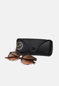 Ochelari de soare Ray-Ban cu lentile brune cu gradient, rame din plastic de culoare scoică închisă, însoțiți de un toc din piele neagră cu închidere tip clapetă.