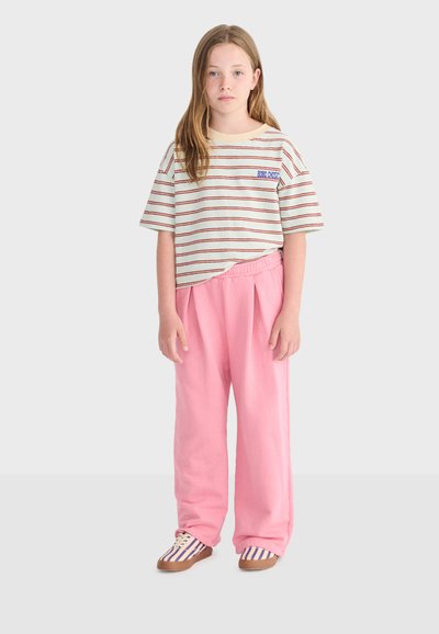 Young girl standing, wearing a cream and red striped short-sleeve shirt, loose pink pants, and striped sneakers on a plain background.
