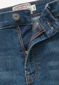Jean en denim bleu foncé avec des coutures cuivres, une fermeture éclair en métal et un bouton pression, portant une étiquette indiquant la taille 34R.