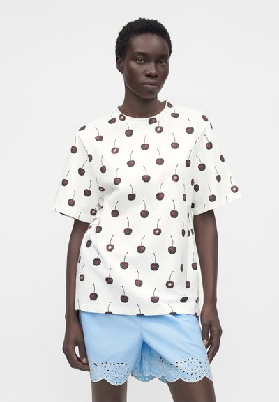 Μοντέλο που φορεί λευκό oversized T-shirt με εκτύπωση κερασιών και ανοιχτό μπλε σορτς με κυματιστό μάτι, πάνω σε απλό φόντο.