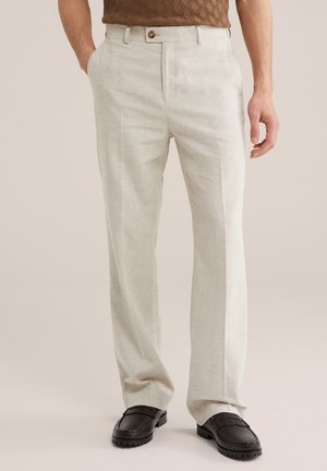 Vīrietis, kurš valkā bēša taisnas bikses ar priekšpusi ar pogu un rāvējslēdzēju, saskaņotas ar melnām ādas loafer apaviem un brūnu teksturētu kreklu.