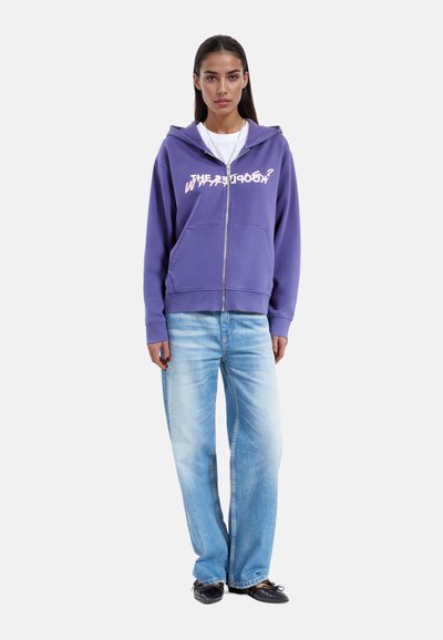 Felpa zip-up viola con tasca frontale e testo stampato bianco, abbinata a jeans a gamba larga blu chiaro e scarpe slip-on nere.
