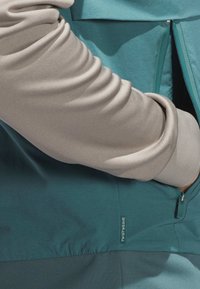 T-shirt à manches longues en tissu beige clair avec une texture lisse, associé à une section unie en bleu sarcelle. Comprend une poche zippée.