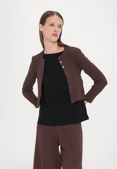 Cardigan côtelé marron avec un col rond, présentant des fermetures à boutons sur le devant. Porté sur un haut noir et un pantalon plissé marron.