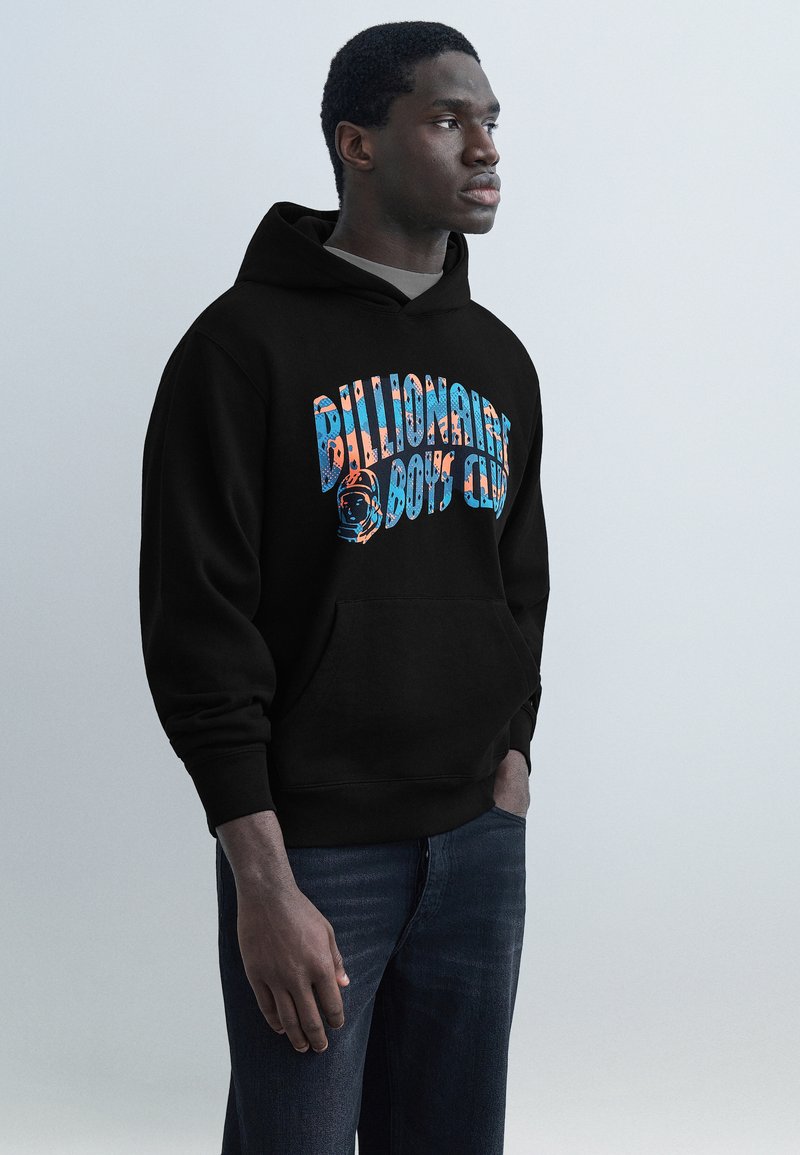 Νέος άνδρας που φοράει μαύρο φούτερ με κουκούλα με πολύχρωμο κείμενο "Billionaire Boys Club" και γραφικό αστροναύτη, στέκεται μπροστά σε απλό φωτεινό φόντο.