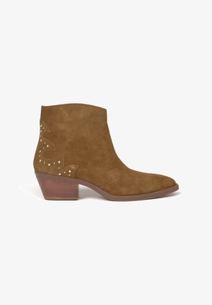 Botines de ante marrón con punta afilada y tacón bajo apilado, que presentan costuras decorativas y tachuelas metálicas en el lateral.