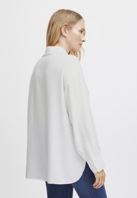 Camicia bianca a maniche lunghe con colletto, texture liscia e polsini con bottoni. Il taglio è rilassato, con un orlo posteriore leggermente più lungo.