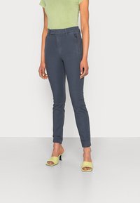 Calças ajuste azul escuro com um bolso com fecho, corte slim e bainhas enroladas, combinadas com uma t-shirt verde-claro e sapatos de salto abertos amarelos.