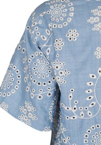 Blouse brodée bleu clair ornée de motifs floraux blancs et de motifs circulaires, avec des manches courtes et un tissu doux et texturé.