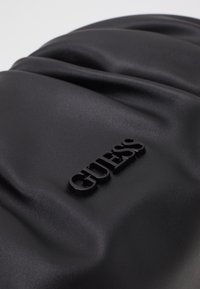 Sort taske i kunstlæder med rynket tekstur og et fremtrædende sort præget "GUESS"-logo på fronten. Glat, mat finish.