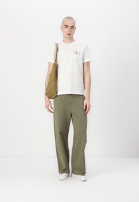 Hvit kortermet t-skjorte med et lite grafisk motiv, kombinert med olivengrønne vidde bukser og en brun tote-bag. Hvite sneakers fullfører antrekket.