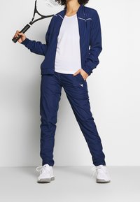 Námornícká sportovní bunda a kalhoty set s bílými akcenty. Hladká, lehká tkanina; model drží tenisovou raketu. Bílé tenisky.