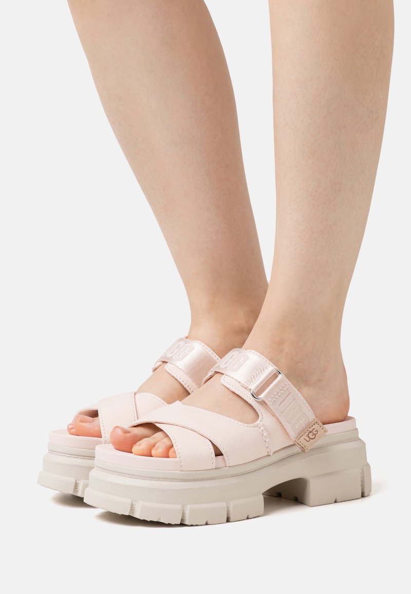 Pieds portant des sandales plateformes épaisses rose pâle avec des sangles croisées épaisses et des sangles réglables à la cheville sur un fond clair.