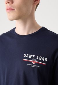 Marineblauwe katoenen t-shirt met de tekst "GANT 1949", een grafisch logo en rood-witte gestreepte accenten op de borst. Standaard crewneck ontwerp.