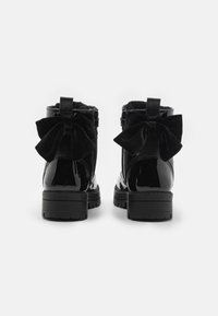 Bottines en cuir verni noir à effet cuir synthétique avec un doublure en peluche noire, présentant un grand nœud en velours à l'arrière et des semelles en caoutchouc durables.