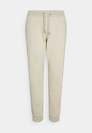 Beige joggingbukser med elastisk taljebånd og snøre foran, med ribbede manchetter og et diskret trykt mønster på højre ben.