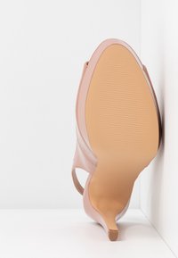 Chaussure à talon haut en cuir verni nude inclinée vers le haut, montrant la semelle lisse et le talon fin sur fond blanc.