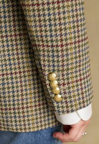 Sako s vzorom houndstooth v odtieňoch svetlohnedej, zelenej, modrej a červenej. Má zlaté gombíky s kvetinovým vzorom na rukávoch. Textúrovaná vlnená tkanina.