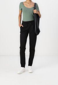 Svarta smala byxor med elastisk midja och sidofickor, i kombination med en formsydd grön topp. Modellen bär en svart väska och vita sneakers.