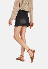 Sandali marroni con design floreale a intaglio, suola piatta e cinturino regolabile alla caviglia, abbinati a una gonna di jeans nera e a un maglione beige lavorato a maglia.