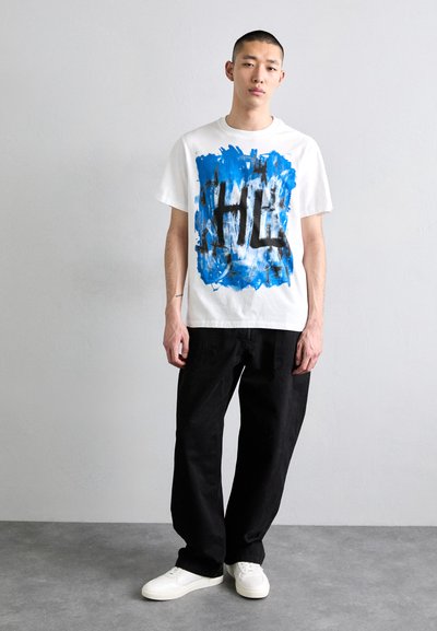 Weißes T-Shirt mit einem blau-schwarzen abstrakten Grafikdesign, kombiniert mit lockeren schwarzen Hosen und weißen Sneakers. Neutraler Hintergrund.