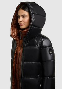 Sort pufferjakke med en skinnende finish, featuring en stor hætte, brun indvendig foring, lynlåslukning og logo på Ærmet.