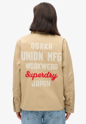 Beige werkjacket met geborduurde teksten inclusief "OSAKA," "UNION MFG," en "Superdry JAPAN" in het wit en rood. Gladde stof.